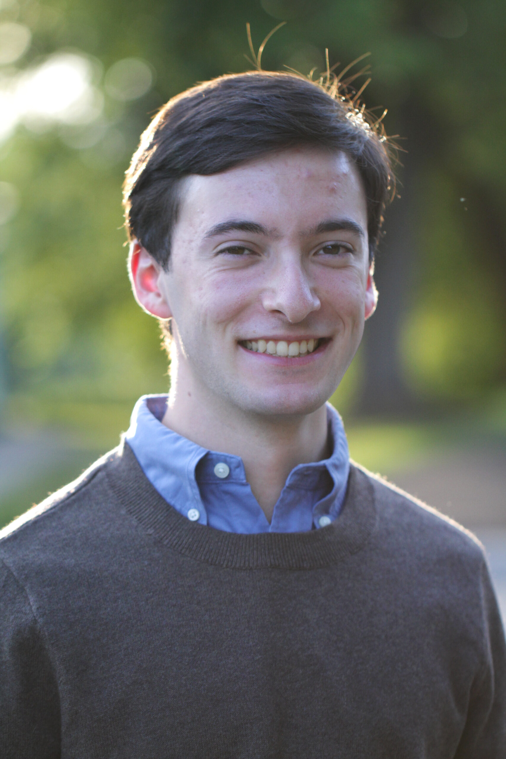 A portrait photo of Xander taken outside with a blurry background. Xander is wearing a brown sweater over a blue button-down shirt. 